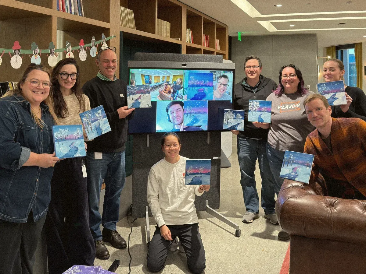 The design team holding up their finished winter paintings from the holiday paint-along, with remote teammates on the screen behind them